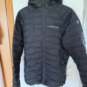 Svart dunjacka Peak Performance - Svart dunjacka från Peak Performance med huva och dragkedja framtill. Jackan har quiltad design, två sidofickor med dragkedja och logga på bröstet. Perfekt för kyliga dagar och har en sportig look. OBS lagad på ena armbågen!!