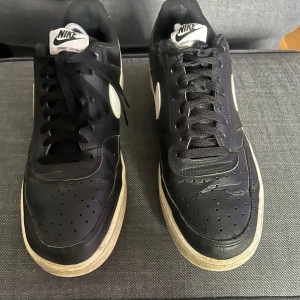 Nike Court Vision Low svart/vit 44 - Svarta Nike Court Vision Low sneakers med vita detaljer och klassisk swoosh på sidan. Skorna har snörning, perforerad tå och platt sula. Tillverkade i skinn och syntet, med retroinspirerad design och tydlig Nike-logga på hälen.