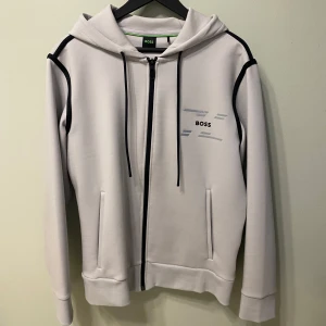 Beige hoodie med dragkedja från HUGO BOSS  - Snygg beige hoodie från HUGO BOSS med svarta detaljer längs axlarna och dragkedja framtill. Tröjan har huva med snörning, två fickor och broderade grå detaljer på bröstet. Perfekt för en chill och stilren look.             Använd fåtal gånger.        Nypris 2800 