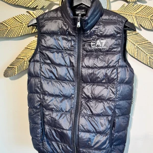 Svart dunväst från EA7 Emporio Armani - Gilet Piumino Unisex stlk 14A (160cm) Snygg mörkblå dunväst från EA7 Emporio Armani med quiltad design och hög krage. Västen har dragkedja framtill och två sidofickor. Perfekt att slänga över en hoodie för en chill look. Logga på bröstet för extra stilpoäng.