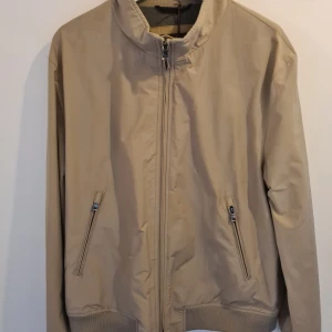 Beige bomberjacka från Oscar Jacobson - Stilren beige bomberjacka från Oscar Jacobson med dragkedja framtill, ribbade muddar och två snedställda fickor med dragkedja. Jackan har en grå insida med innerficka och klassisk bomberkrage. Perfekt för dig som vill ha en clean och tidlös look. Oanvänd. Nypris 2999kr. 