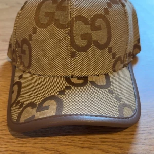 Gucci beige keps med GG-mönster - Snygg beige keps från Gucci med det klassiska GG-mönstret i brunt över hela kepsen. Kepsen har böjd skärm med brunt kantband och är tillverkad i bomull och polyester. Perfekt för dig som vill ha en lyxig och trendig accessoar.