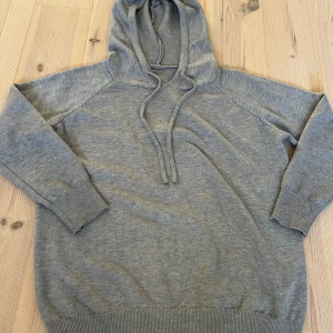 Grå kashmir hoodie - En enkel och stilren grå kashmir hoodie med huva och snörning framtill. Tröjan har långa ärmar och breda ribbade muddar vid ärmslut och nederkant. Perfekt för chill dagar och passar till det mesta. Materialet känns mjukt och är väldigt skönt enligt mig. Passar nog bäst på 164-170cm. 
