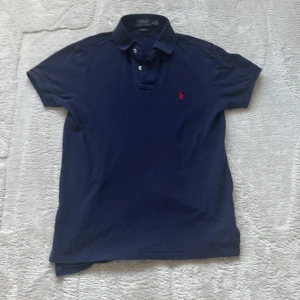 Marinblå pikétröja från Polo Ralph Lauren - Klassisk marinblå pikétröja från Polo Ralph Lauren med röd broderad logga på bröstet. Tröjan har krage, knäppning med två knappar och är tillverkad i bomull. Custom fit-modell som sitter snyggt och passar till många stilar.