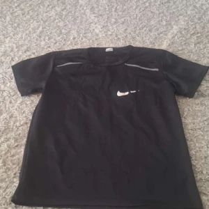 Svart Nike t-shirt med tryck - Svart t-shirt från Nike med klassisk vit swoosh-logga på bröstet och diskreta grå detaljer vid axlarna. Modellen har rund hals och korta ärmar, perfekt för dig som gillar sportig stil. Materialet är mjukt och lätt polyester.