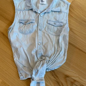Ljusblå ärmlös jeansblus H&M - Säljer en ljusblå ärmlös jeansblus från H&M med två bröstfickor, knappar framtill och knytning nedtill. Perfekt för en avslappnad och cool look. Materialet är mjukt denim och toppen har klassisk skjortkrage. Det finns en fläck (se bild 2), men den är inte utmärkande. 