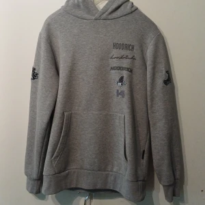 Grå hoodie från Hoodrich  - Säljer en grå hoodie från Hoodrich i storlek M. Tröjan har tryck och broderade detaljer både fram och bak samt på huvan. Klassisk känguruficka och ribbade muddar. Perfekt för dig som gillar streetwear och vill ha en snygg och bekväm look.