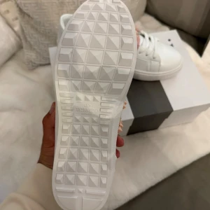 Valentino Rockstud sneakers vit/rosa - Snygga Valentino Garavani Rockstud sneakers i vitt skinn med rosa band och ikoniska nitar. Klassisk låg modell med snörning och rund tå. Perfekt för dig som vill sticka ut med exklusiva detaljer och modern design.