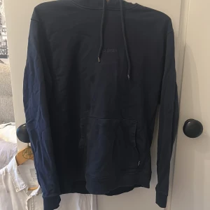 Mörkblå hoodie från Les Deux - Snygg mörkblå hoodie från Les Deux med klassisk känguruficka och dragsko i huvan. Tröjan har långa ärmar och ribbade muddar. Perfekt för en chill och stilren look.