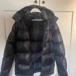 Svart pufferjacka från Polo Ralph Lauren - .En riktigt snygg, varm vinter jacka! Köptes vintern 2024 användes endast den vintern och inte sedan dess. Jackan är som ny men har några små fläckar liknande lim fläckar på baksidan av vänster arm och på framsidan på vänster sida på jackan. Inga fläckar som syns mycket då de smälter in i glanset på jackan! Annars är jackan i topp skick! Pris går att diskuteras