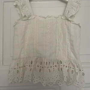 Vit broderad blus med volang - Supersöt vit blus med breda axelband och broderade detaljer. Toppen har volangkant nertill och spetsmönster längs hela plagget. Axelbanden är dekorerade med hålbroderi och den här är asfin att ha både till jeans eller till finkjol! Säljer för 179, nypris 250 💗