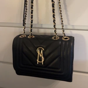 Svart quiltad handväska från Michael Kors - Snygg svart quiltad handväska från Michael Kors med guldfärgade kedjor och logga framtill. Väskan har en stilren design med kedjeaxelrem och elegant struktur. Perfekt för dig som vill ha en trendig accessoar med lyxig känsla.