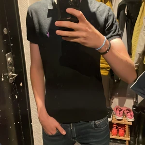 Svart pikétröja från Ralph Lauren - Svart pikétröja från Ralph Lauren med klassisk krage och korta ärmar. Tröjan har en broderad rosa logga på bröstet och är i bomull. Snygg och stilren modell som sitter slim.