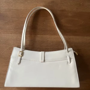 Gräddvit axelväska med smala remmar - Snygg vit axelväska med stilren, rektangulär form och smala axelremmar. Väskan har guldfärgade detaljer och dragkedja upptill. Tillverkad i slätt skinnmaterial och har flera fack inuti för smart organisering. Perfekt för dig som gillar minimalistisk och clean stil.