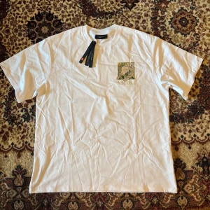 Vit Amiri t-shirt med leopardtryck - Vit t-shirt från Amiri i bomull med coolt leopardtryck och gröna detaljer både fram och bak. Klassisk passform och rund halsringning. Stort Amiri-tryck under motivet. Perfekt statement-plagg för dig som gillar streetstyle.