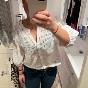 Vit blus med broderade hål och puffärm - Säljer en vit blus i bomull med broderade hål och puffiga trekvartsärmar. Blusen har knappar framtill, rundad halsringning och är croppad i modellen. Perfekt för dig som gillar romantiska och luftiga plagg med snygga detaljer.