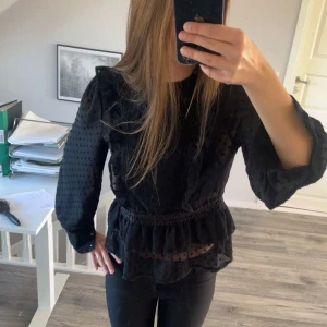 Svart blus med volang - Säljer en svart blus med volangdetaljer och diskret prickigt mönster. Blusen har lång ärm, rund hals och markerad midja med spetskant. Ärmarna går även att knäppa om man vill. Storlek 38 men passar även 36/S. 