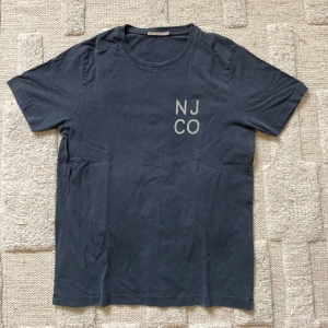 Mörkblå t-shirt från Nudie Jeans Co - Snygg mörkblå t-shirt från Nudie Jeans Co med trycket 'NJ CO' på bröstet. Klassisk rund halsringning och korta ärmar. Tillverkad i mjuk bomull som känns skön mot huden. Perfekt för en avslappnad och stilren look.
