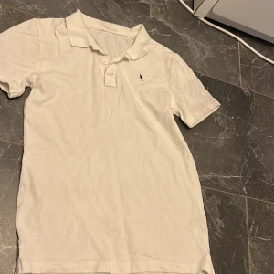 Vit pikétröja från H&M med brodyr - Vit pikétröja från H&M i bomull med klassisk krage och knäppning framtill. Tröjan har korta ärmar och en liten svart broderad logga på bröstet. Perfekt basic till garderoben och enkel att matcha med allt.