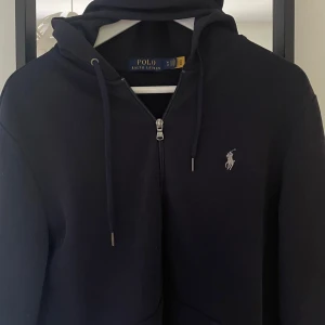 Mörkblå hoodie från Polo Ralph Lauren - Snygg mörkblå hoodie från Polo Ralph Lauren med dragkedja hela vägen, huva med snörning och klassisk logga broderad på bröstet. Tillverkad i mjuk bomullsmix som känns riktigt skön. Perfekt för chill dagar eller när du vill ha en clean look.
