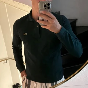 Mörkgrön pikétröja från Lacoste - Snygg mörkgrön långärmad pikétröja från Lacoste med klassisk krokodillogga på bröstet. Tröjan har krage och knappar framtill, tillverkad i mjuk bomull med normal passform. Perfekt för dig som gillar stilrena och sportiga plagg.