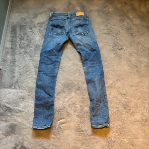 Blå Nudie Jeans Skinny Fit - Säljer ett par klassiska blå jeans från Nudie Jeans med skinny fit. Jeansen har snygga slitningar, kontrastsömmar och Nudie-logga i midjan. Perfekt för dig som gillar skinny/slim jenas. Storlek: W:25, L:32