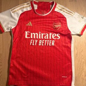 Arsenal hemmatröja Adidas röd/vit - Säljer en Arsenal hemmatröja från Adidas i rött och vitt med guldiga detaljer. Tröjan har korta ärmar, blixtmönster och klubbmärke på bröstet. Materialet är lätt och andas, perfekt för match eller träning. Emirates Fly Better tryck på framsidan.