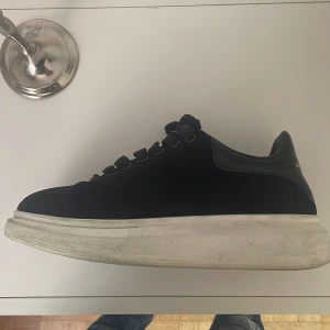 Alexander McQueen svarta sneakers - Säljer ett par svarta Alexander McQueen sneakers med chunky vit sula och klassisk logga bak på hälen. Skorna har rund tå, breda svarta skosnören och är tillverkade i skinn för en lyxig känsla. Perfekta för dig som gillar statement sneakers med stilren design.