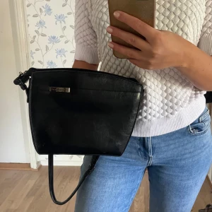 Svart axelväska från Puccini i skinn - Snygg svart axelväska från Puccini i äkta skinn med stilren, fyrkantig form. Väskan har ett diskret metallmärke framtill och justerbar axelrem. Perfekt storlek för det viktigaste och stängs med dragkedja upptill.