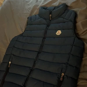 Mörkblå dunväst från Moncler - Snygg mörkblå dunväst från Moncler med klassisk quiltad design och hög krage. Västen har dragkedja framtill, två fickor med dragkedja och Moncler-logga på bröstet. Perfekt för lager-på-lager och streetstyle.Pris ej hugget!