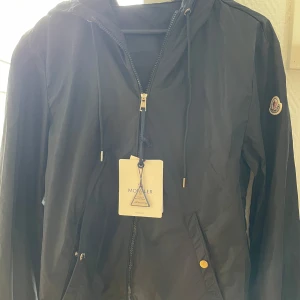 Svart vindjacka från Moncler - Svart vindjacka från Moncler med huva och dragsko, klassisk logga på ärmen och snygga detaljer i silver. Jackan har dragkedja framtill och fickor med tryckknappar. 