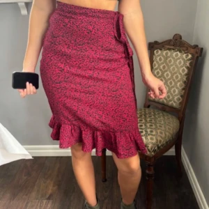 Röd leopardmönstrad kjol med volang - Snygg röd kjol med svart leopardmönster och volangkant nertill. Kjolen har omlottkänsla med knyt i sidan och en dekorativ volang som löper baktill. Perfekt för dig som vill sticka ut med en trendig look.