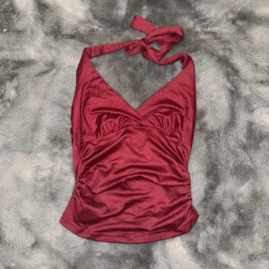 Vinröd halterneck topp med knyt - Snygg vinröd halterneck topp med knyt i nacken och v-ringad front. Toppens material har en lätt glans och smickrande rynkade detaljer framtill. Perfekt för dig som vill sticka ut med en elegant och trendig look.