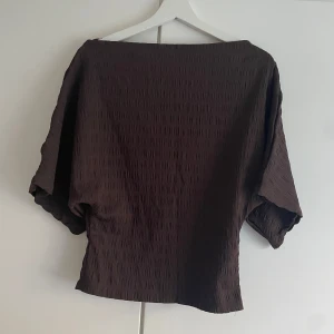Brun topp från Nelly med struktur - Mörkbrun topp från Nelly i storlek S med lös passform och korta ärmar. Tyget har en snygg strukturerad yta som ger en cool look. Perfekt att matcha med byxor i samma material för en trendig outfit.