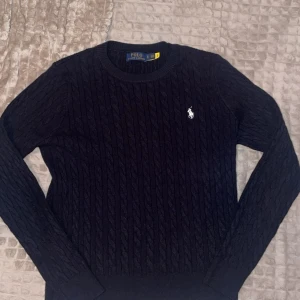 Polo Ralph Lauren Tröja - Kabelstickad mörkblå tröja från Polo Ralph Lauren med klassisk rund halsringning och broderad vit logga på bröstet. Långärmad modell i mjuk bomull, perfekt för lager-på-lager under höst och vinter. Den är rätt så kort vid midjan annars perfekt! Den är M men sitter som S
