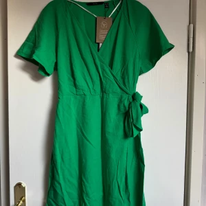 Grön omlottklänning med knyt - Snygg grön omlottklänning från Veromoda med korta ärmar och knyt i midjan. Klänningen har v-ringning och är tillverkad i ett mjukt tyg. Perfekt för en fräsch och färgstark look under varmare dagar.