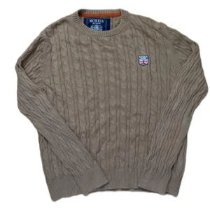 Beige kabelstickad tröja från Morris - Kabelstickad beige tröja från Morris med rund halsringning och broderad patch med brittisk flagga på bröstet. Tröjan har ribbade muddar och är tillverkad i mjukt material som passar perfekt för kyligare dagar.