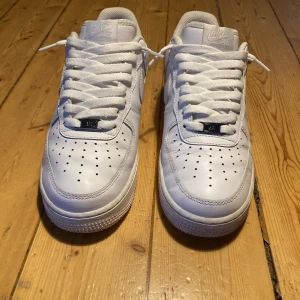 Nike Air Force 1 vita  - Klassiska Nike Air Force 1 sneakers i helvitt skinn med perforerad tå, rund tå och platt sula. Swoosh-logga på sidorna och broderad Nike Air-text på hälen. Tjocka vita snören och låg profil gör dem till en ikonisk streetwear-sko.