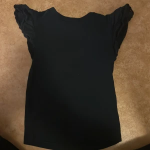 Svart volangärmad topp Gina Tricot - Svart topp från Gina Tricot med snygga volangärmar som ger en cool och feminin touch. Modellen är enkel och rak med rund halsringning. Perfekt att styla med jeans eller kjol för en trendig look.