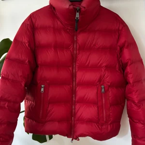 Röd dunjacka från Polo Ralph Lauren - Snygg röd dunjacka från Polo Ralph Lauren med hög krage och två dragkedjeförsedda fickor. Jackan har quiltad design och klassisk logga broderad på bröstet.. Knappt använd, då den är lite för stor för mig. Priser går att diskutera 