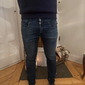 Blå Replay jeans (Anbass) - Säljer ett par blå jeans från Replay med klassisk femficksdesign och dragkedja. Jeansen har en smal passform och snygga slitningar för en cool look. Märkespatch bak i läder och Replay-logga vid fickan. Perfekta för dig som gillar stilrena och trendiga jeans.