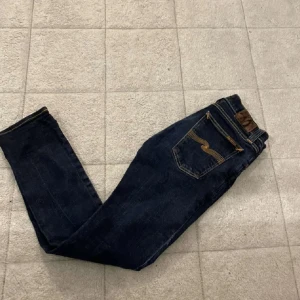 Mörkblå jeans från Nudie Jeans - Snygga mörkblå jeans från Nudie Jeans med klassisk femficksdesign och kontrastsömmar på bakfickan. Jeansen har en rak passform och är tillverkade i slitstarkt denimtyg. Perfekta för dig som gillar stilrena och tidlösa jeans.