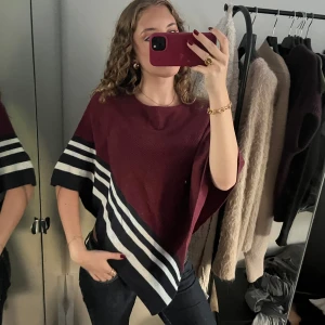 Vinröd stickad ponchotröja Holly & Whyte - Ponchotröja i vinrött från Holly & Whyte med breda vita och mörkblå ränder längs ärmar och nederkant. Stickad i mjukt material, har dekorativa knappar på ena axeln och loose passform. Perfekt för lager-på-lager under höst och vinter. Passar storlekar mellan xs-L