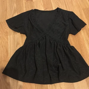 Svart blus med v-ringning och spets - Svart blus med v-ringning och fina spetsdetaljer. Blusen har korta ärmar och ett omlottliknande liv, samt broderade hålmönster över hela tyget. Den har en luftig och lätt utsvängd passform som ger en söt och trendig look.