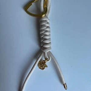 Nyckelring - Unikt armband med vit flätad snodd, guldfärgad hjärtformad detalj och en liten -berlock i guld. Perfekt accessoar för dig som gillar statement-smycken och exklusiv känsla.