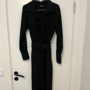 Svart långärmad jumpsuit med bälte - Stilren svart långärmad jumpsuit från Bik Bok med klassisk krage, knappar framtill och markerat midjebälte. Perfekt för dig som gillar en enkel men snygg look. Klänningen har fickor på bröstet och är i ett mjukt, följsamt material.