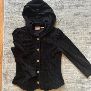 Svart velourkofta Juicy Couture S - Svart kofta från Juicy Couture i mjuk velour med diskret mönster, guldiga knappar och huva. Långärmad och figurnära modell, perfekt för en chill och snygg look. Klassisk logga på insidan och ribbade muddar vid ärmslut. Vintage 