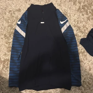 Nike Dri-Fit träningströja blå slim fit - Nike Dri-Fit långärmad träningströja i slim fit med marinblå kropp och blåmönstrade ärmar. Snygga vita detaljer och dragkedja framtill. Perfekt för dig som vill ha en sportig och stilren look på gymmet eller ute.
