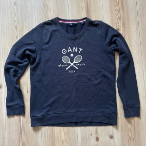 Mörkblå sweatshirt från Gant - M - Mörkblå sweatshirt från Gant med tryck av två tennisrack och texten 'Båstad Sweden 2016' på bröstet. Tröjan har rund halsringning, långa ärmar och ribbade muddar. Perfekt för en sportig och avslappnad stil.