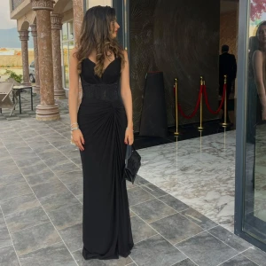 Svart långklänning med drapering - Supersnygg svart långklänning med v-ringad omlottdesign och markerad midja. Klänningen har draperad detalj i sidan och smala axelband. Perfekt för bal eller festliga tillfällen. Materialet är mjukt och följsamt, vilket ger en elegant siluett.